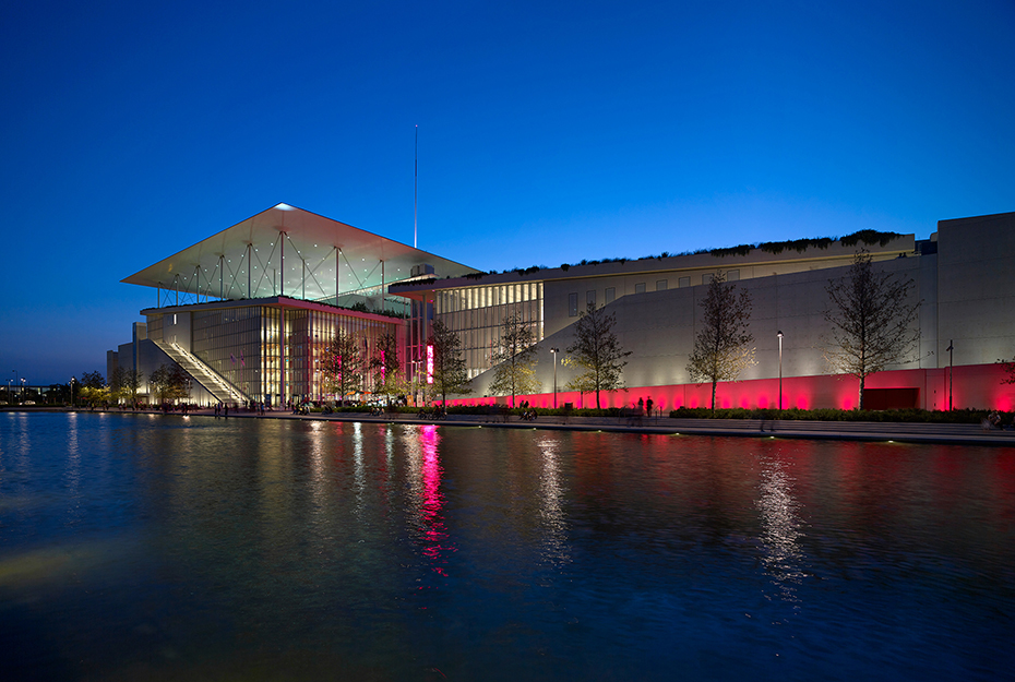 Φωτογραφία που δείχνει μια λίμνη στο τέλος της οποίας υπάρχει ένας τοίχος που φωτίζεται και την άκρη φαίνεται ένα μεγάλο γυάλινο κτήριο 