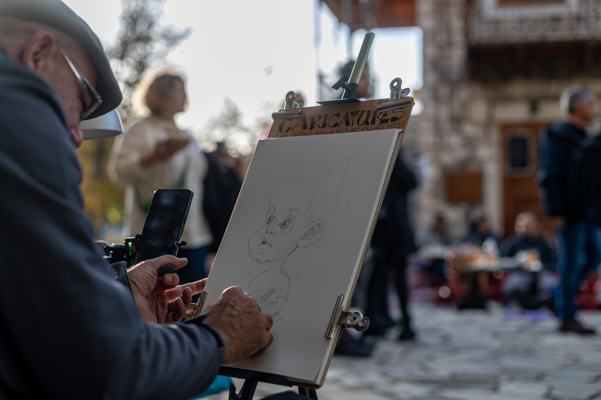 Ένας άντρας στέκεται στην πλατεία του χωριού και ζωγραφίζει ένα μικρό παιδάκι με τη μορφή καρικατούρας. 