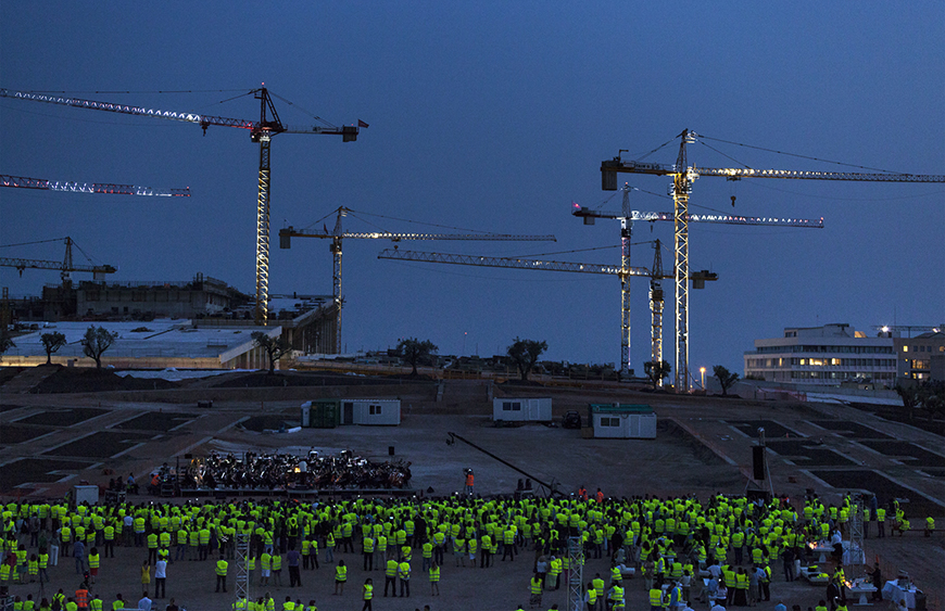 Πλήθος ανθρώπων μέσα σε ένα εργοτάκιο φοράει το ίδιο γιλέκο εργασίας και έξι γερανοί τοποθετημένοι στον χώρο