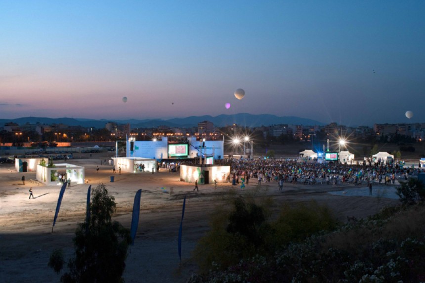 Μακρινή φωτογραφία τραβηγμένη τη νύχτα η οποία δείχνει πλήθος κόσμου σε ένα εργοτάξιο να κοιτούν προς τη σκηνή 
