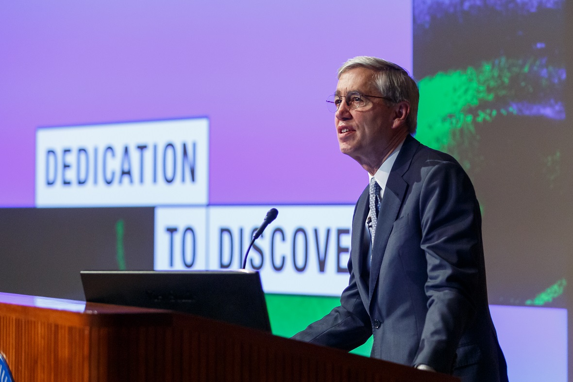 Φωτογραφία ενός άντρα να μιλάει στο βήμα και δίπλα του να προβάλλεται μια φωτογραφία η οποία γράφει DEDICATION TO DISCOVERY 