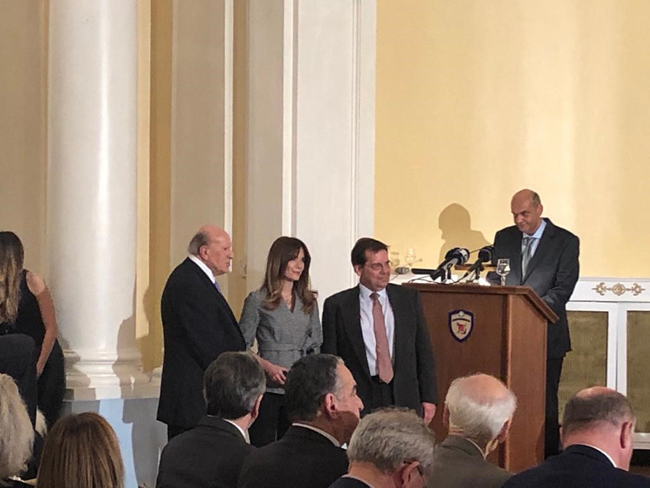 Η Άννα-Κύνθια Μπουσδούκου στέκεται ανάμεσα σε δύο άνδρες μπροστά σε ένα καθισμένο κοινό μετά την αποδοχή του Βραβείου Δημοσιογραφίας και Τεχνολογίας «Γιώργος Καραγιάννης»