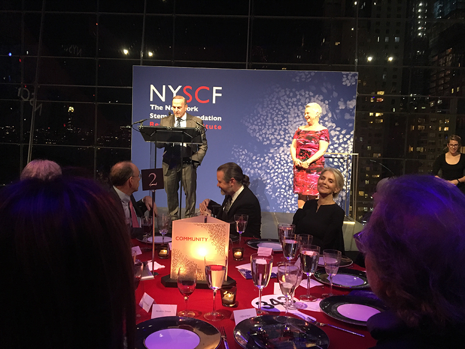 Κόσμος κάθεται σε ένα στρογγυλό τραπέζι και παρακολουθεί τον άντρα που βγάζει λόγο στο βήμα πάνω στη σκηνή και τη γυναίκα που είναι δίπλα του στη σκηνή και γελάει 