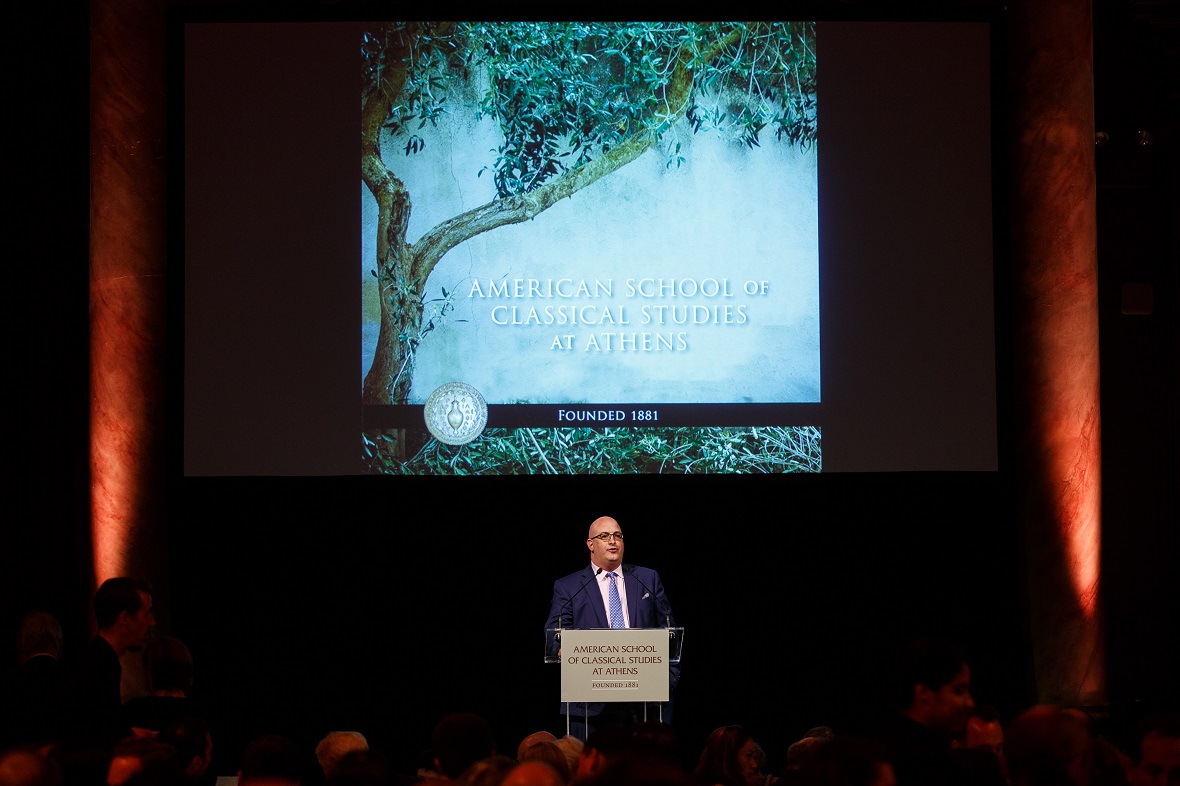 Ένας άνδρας δίνει λόγο στη σκηνή μπροστά από μια οθόνη που γράφει 