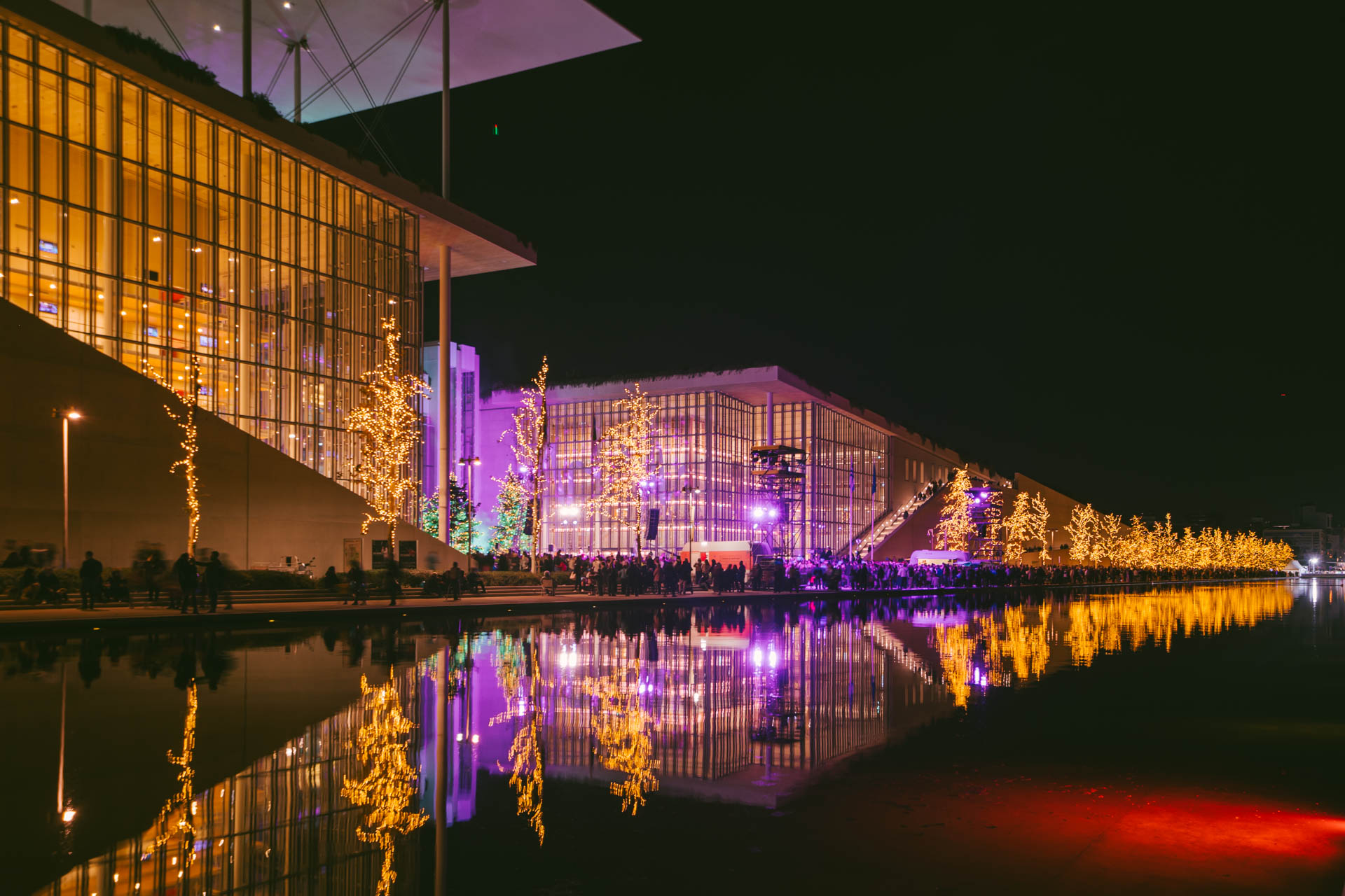Το Κέντρο Πολιτισμού Ίδρυμα Σταύρος Νιάρχος, φωτισμένο με μωβ και χρυσό, ενώ το πλήθος κόσμου και τα φωτισμένα πλατάνια καθρεφτίζονται στο Κανάλι.  