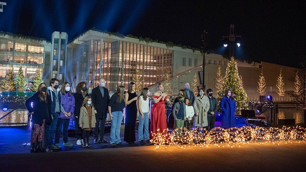 Άνθρωποι και παιδιά, μερικοί κρατώντας μικρόφωνα, στέκονται μπροστά από το φωτισμένο Κέντρο Πολιτισμού Ίδρυμα Σταύρος Νιάρχος