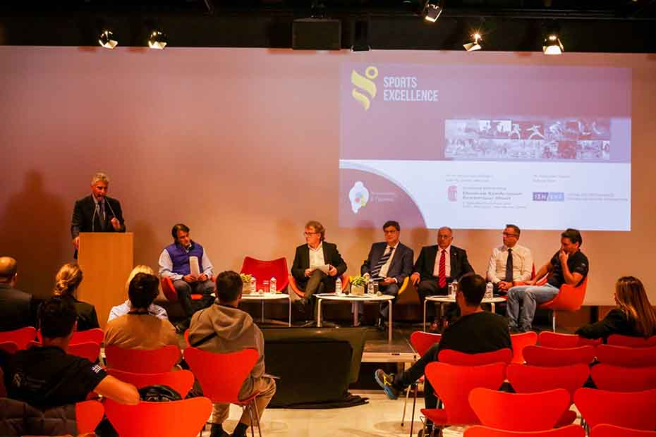 Μακρινή φωτογραφία που δείχνει το κοινό να κάθεται σε καρέκλες και να παρακολουθεί τους άντρες που βρίσκονται στη σκηνή την ώρα που ο ένας μιλάει στο βήμα