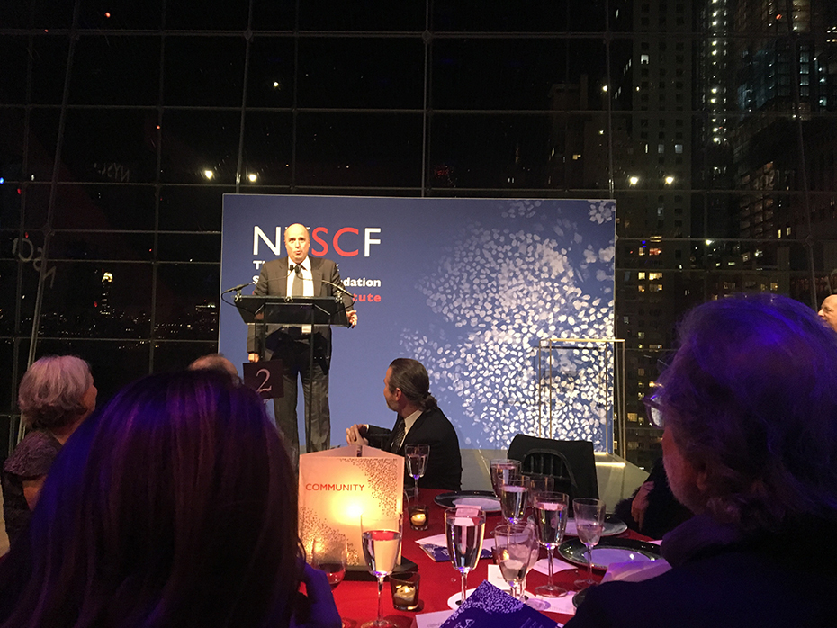 Ο Αντρέας Δρακόπουλος, Πρόεδρος του Ιδρύματος Σταύρος Νιάρχος, μιλάει στο βήμα του NYSCF και ο κόσμος από κάτω κάθεται σε τραπέζια και τον παρακολουθεί 