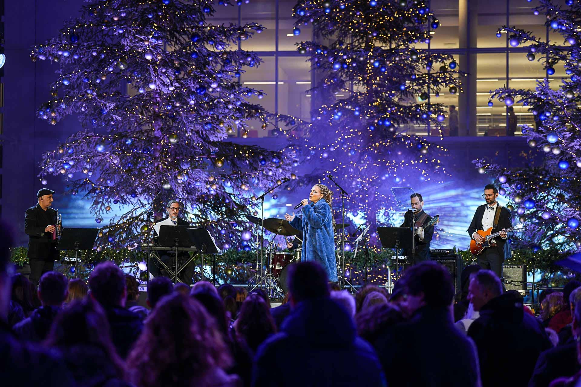 Μια ομάδα μουσικών στη σκηνή, με μεγάλα χριστουγεννιάτικα δέντρα στο φόντο.