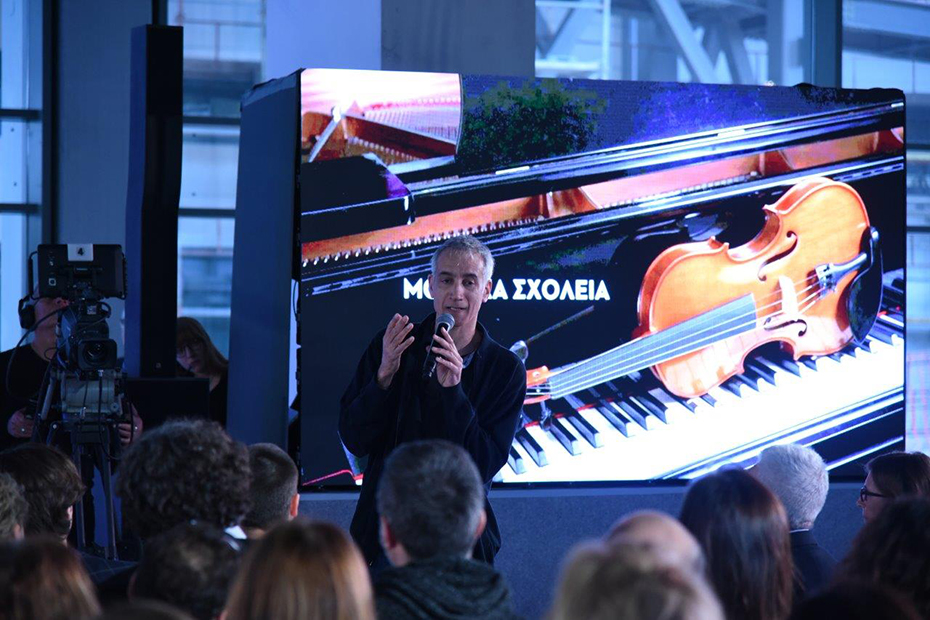 Ένας άντρας είναι όρθιος στη σκηνή και βγάζει λόγο κρατώντας ένα μικρόφωνο ενώ πίσω τους υπάρχει ένα φόντο το οποίο απεικονίζει ένα βιολί να είναι τοποθετημένο στα πλήκτρα ενός πιάνου 