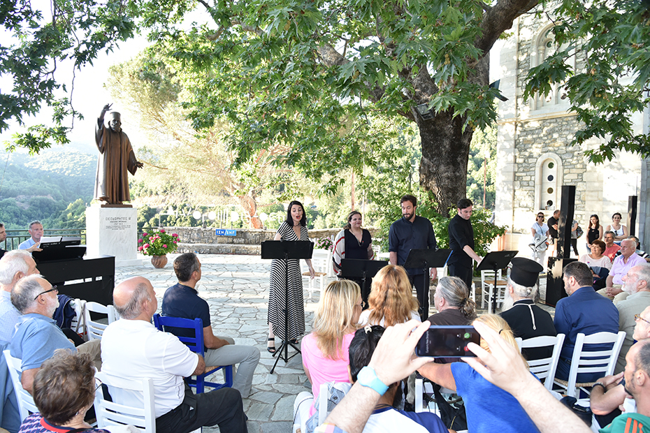 Πλήθος ανθρώπων κάθονται στην κεντρική πλατεία του χωριού και παρακολουθούν τέσσερις καλλιτέχνες 