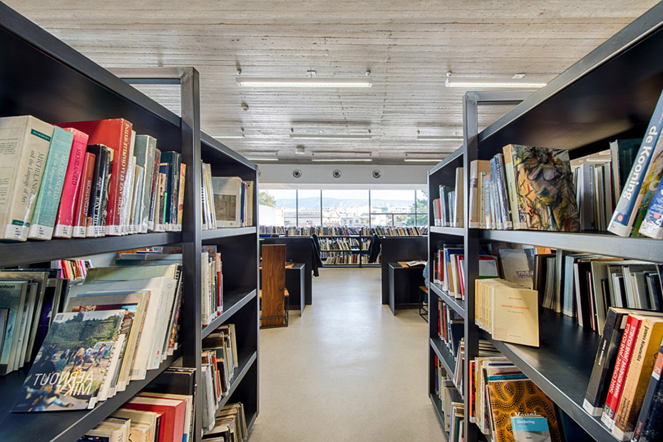 Φωτογραφία τραβηγμένη ανάμεσα από δύο ράφια γεμάτα με βιβλίαπου δείχνει τον υπόλοιπο χώρο της βιβλιοθήκης