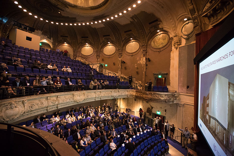 Φωτογραφία που δείχνει την κεντρική σκηνή ενός εμβληματικού θεάτρου στην οποία υπάρχει κοινό το οποίο παρακολουθεί την εικόνα που προβάλλεται, η οποία δείχνει ένα μεγάλο, μοντέρνο κτήριο 