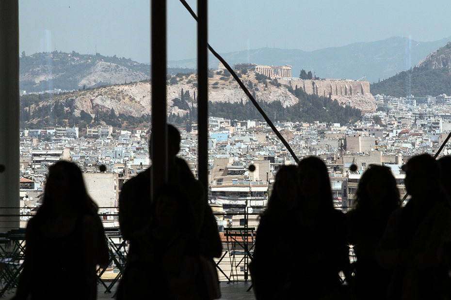 Μια ομάδα ανθρώπων κοιτάζει έξω από ένα παράθυρο με θέα την Ακρόπολη και την Αθήνα.