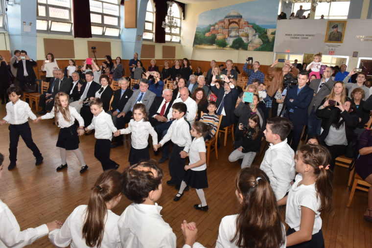 Μικροί μαθητές, κρατιούνται χέρι χέρι σε κύκλο και χορεύουν ελληνικό παραδοσιακό χορό και το κοινό τους παρακολουθεί 