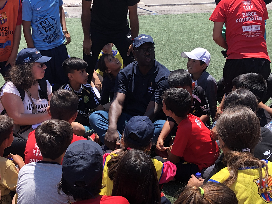 Ένας άντρας και μια ομάδα μικρών παιδιών κάθονται οκλαδόν σε ένα γήπεδο ποσφαίρου και συζητούν 