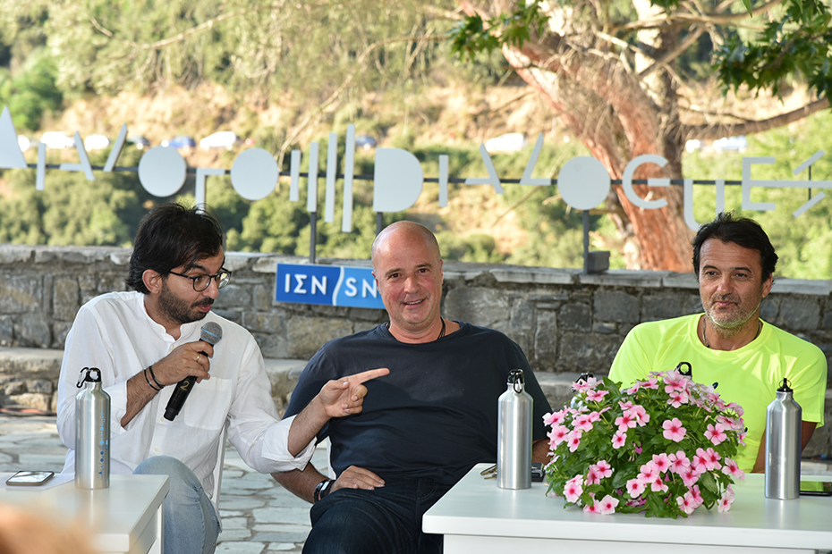 Ο Αντρέας Δρακόπουλος, Πρόεδρος του Ιδρύματος Σταύρος Νιάρχος, κάθεται στην κεντρική πλατεία ενός χωριού και χαμογελά όσο ο άντρας που κάθεται δίπλα του κρατάει το μικρόφωνο και μιλά 