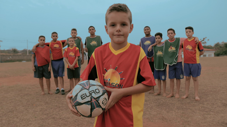 Ένα μικρό αγόρι κρατάει μια μπάλα ποδοσφαίρου και από πίσω του στέκονται αγκαλιασμένα τα υπόλοιπα μέλη της ομάδας 