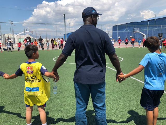 Ένας άντρας και μια ομάδα μικρών αγοριών κρατιούνται χέρι χέρι σχηματίζοντας έναν κύκλο σε ένα γήπεδο ποδοσφαίρου 