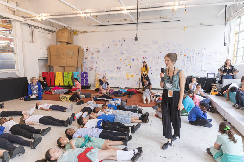 Μια γυναίκα περπατάει και μιλάει σε ένα μικρόφωνο ενώ πολλά παιδιά ξαπλώνουν στο πάτωμα με τα μάτια τους κλειστά