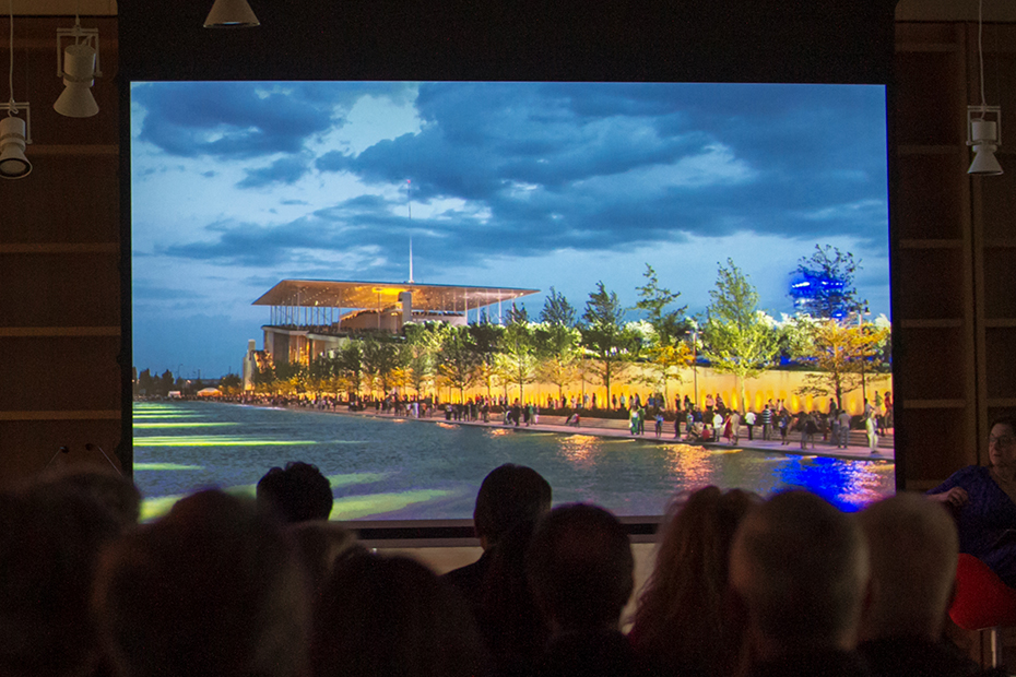 Σε μια οθόνη του σινεμά προβάλλεται ένα μεγάλο γυάλινο κτήριο το οποίο έχει μπροστά του δέντρα και μια λίμνη και ο κόσμος από κάτω κάθεται και παρακολουθεί 