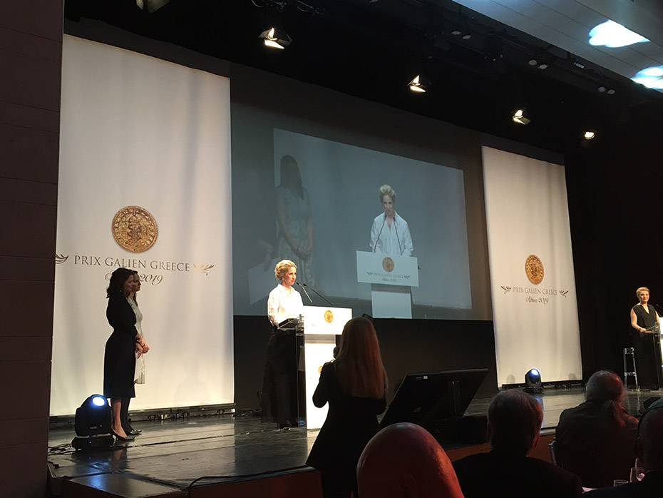 Τέσσερις γυναίκες βρίσκονται στη σκηνή, οι δύο στέκονται στο ένα πλάι της σκηνής, η άλλη στο άλλο και η τέταρτη γυναίκα έιναι στο βήμα και μιλάει και η οθόνη προβάλει τη γυναίκα που μιλάει στο βήμα