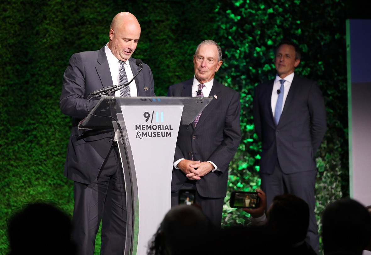 Andreas Dracopoulos, President of the Stavros Niarchos Foundation, at the podium of the 9 I II Memorial & Museum, makes a speech, while two other men stand on stage and watch him