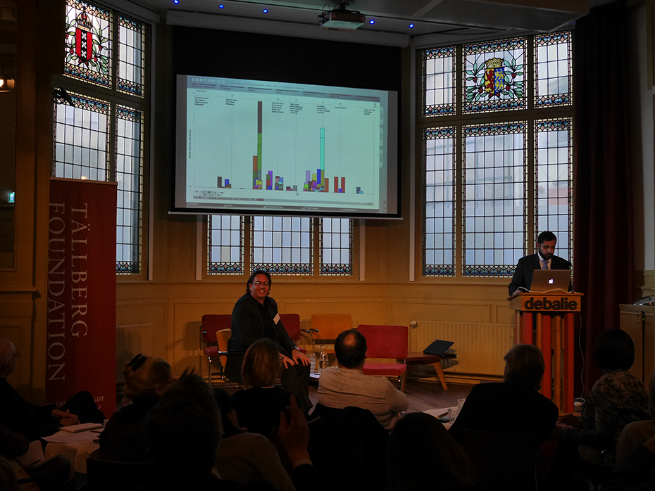 Ένας άνδρας κάθεται μπροστά σε ένα ακροατήριο, ενώ πίσω του εμφανίζεται ένα  διάγραμμα σε μια οθόνη.