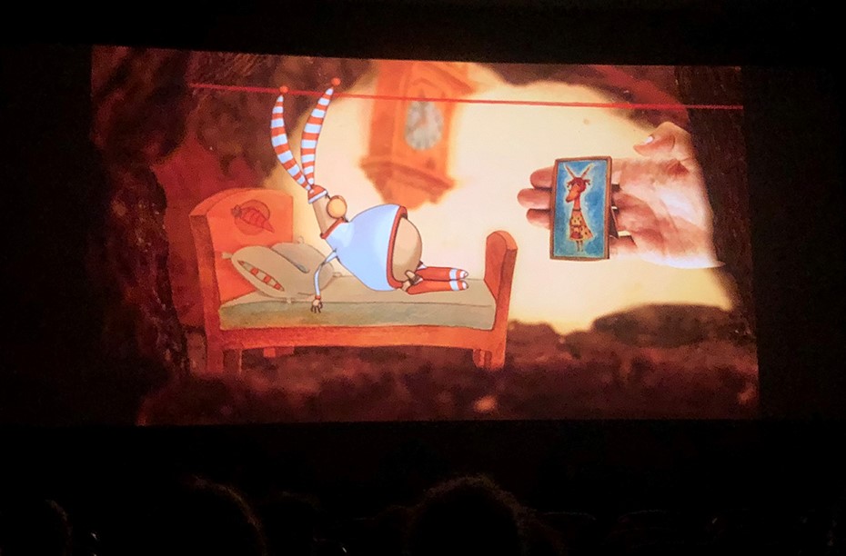 Φωτογραφία στην οποία φαίνεται μια μεγάλη οθόνη του σινεμά στην οποία προβάλλεται ένα κινούμενο σχέδιο να είναι ξαπλωμένο σε ένα κρεβάτι και μια παλάμη να του δείχνει έναν καθρέφτη στον οποίο απεικονίζεται μια καμηλοπάρδαλη 