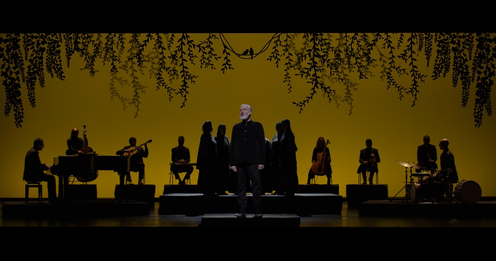 Ένας άνδρας στέκεται στο φως, ενώ μια μπάντα και μια ομάδα γυναικών στέκονται στις σκιές, στην Εναλλακτική Σκηνή της Εθνικής Λυρικής Σκηνής