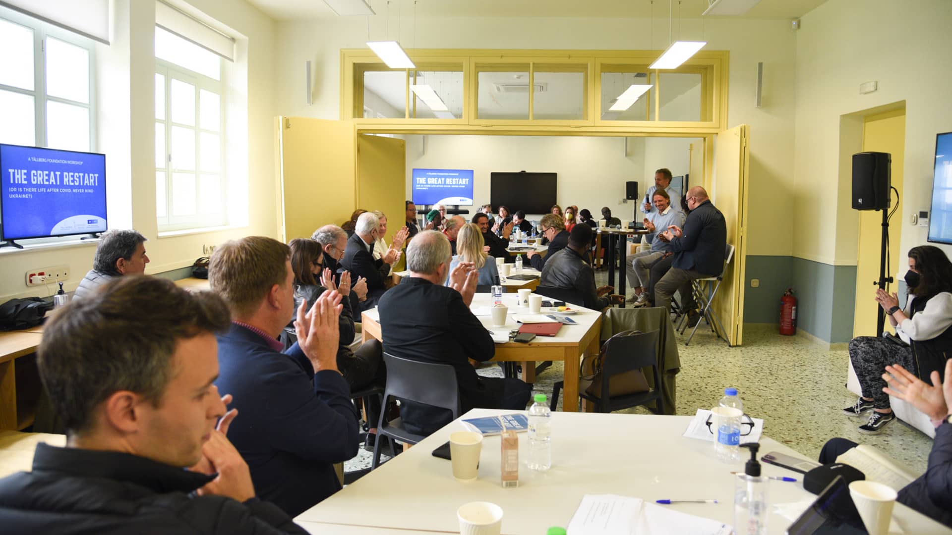 Πολλοί άνθρωποι καθισμένοι σε τραπέζια χειροκροτούν 