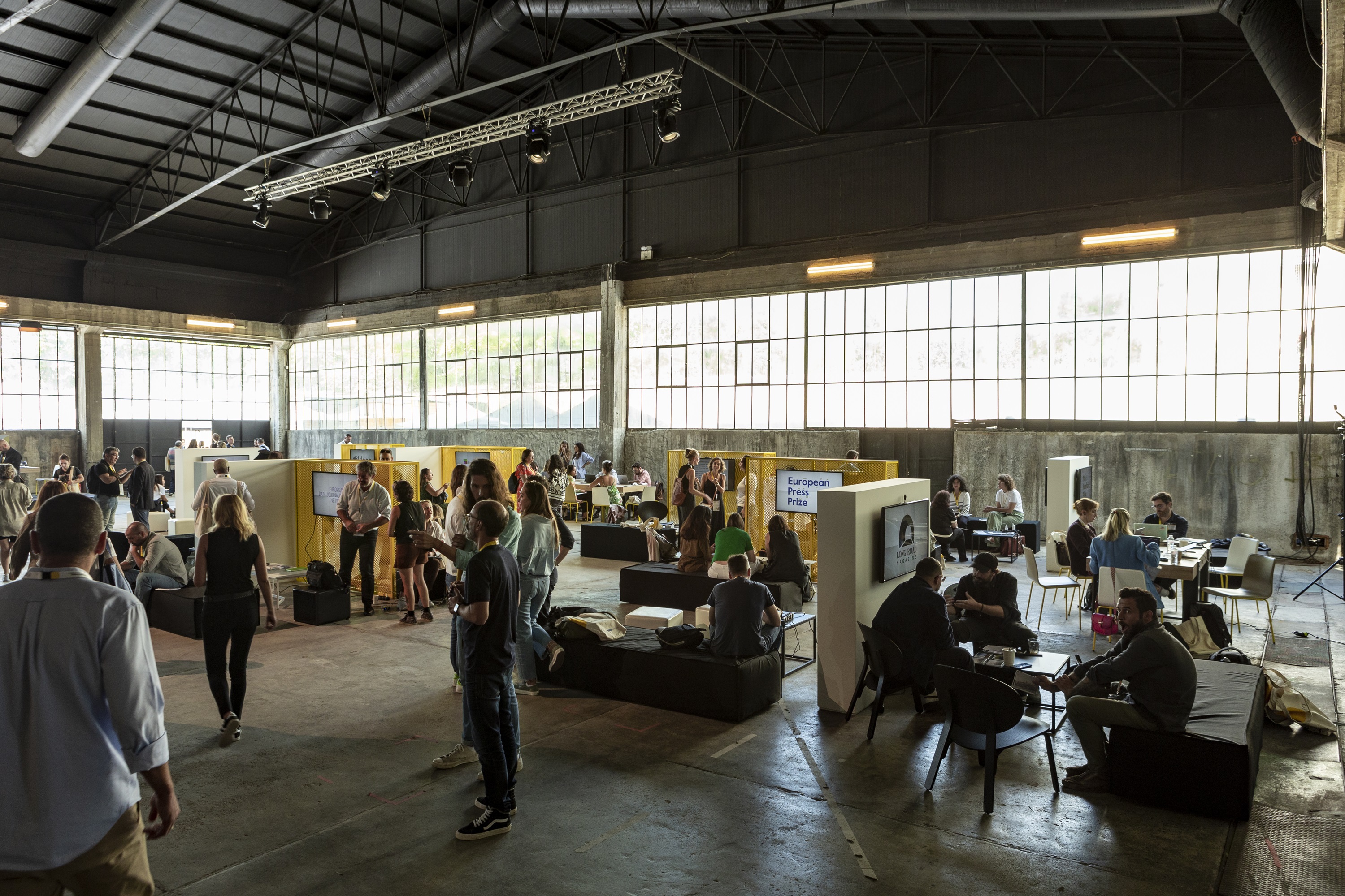 In the interior of an exhibition, there are marble walls scattered around the room on which screens are hung, and around them are chairs and sofas on which a crowd of people sit and talk. 