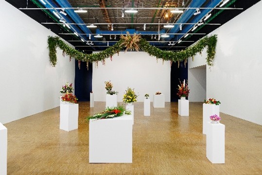 Table white stands, in an art exhibition, on which are bouquets of various colorful flowers as exhibits, while a large garland of green leaves hangs from the ceiling, and in the center of the garland is a bouquet of dried plants, giving the impression of a star.