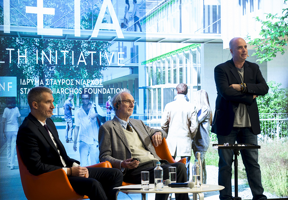 Ο Αντρέας Δρακόπουλος, Πρόεδρος του Ιδρύματος Σταύρος Νιάρχος στέκεται όρθιος στη σκηγή και δίπλα του υπάρχουν δύο άλλοι άντρες καθισμένοι 