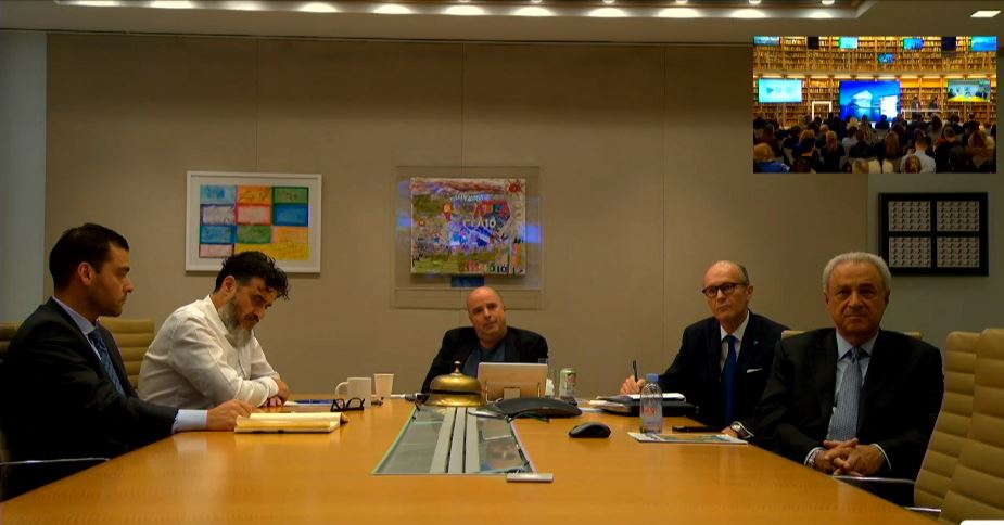 Φωτογραφία μέσα από μια ζωντανή σύνδεση η οποία δείχνει πέντε άντρες να κάθονται γύρω από ένα μεγάλο τρπαέζι συνεδριάσεων και να επικοινωνούν με ένα κοινό το οποίο κάθεται σε καρέκλες