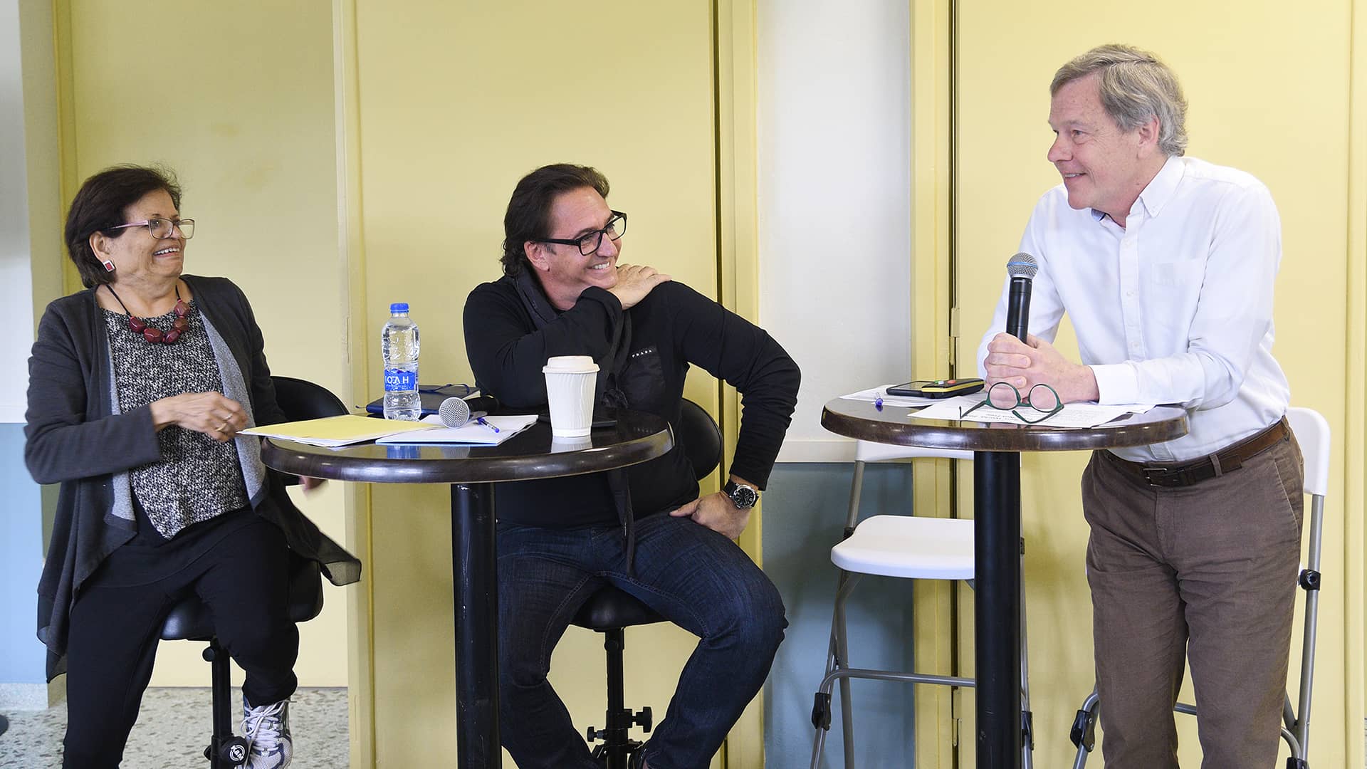 Ένας άντρας και μια γυναίκα κάθονται σε ψηλό τραπέζι και χαμογελούν στον άντρα που κάθεται όρθιος δίπλα τους κρατώντας ένα μικρόφωνο 