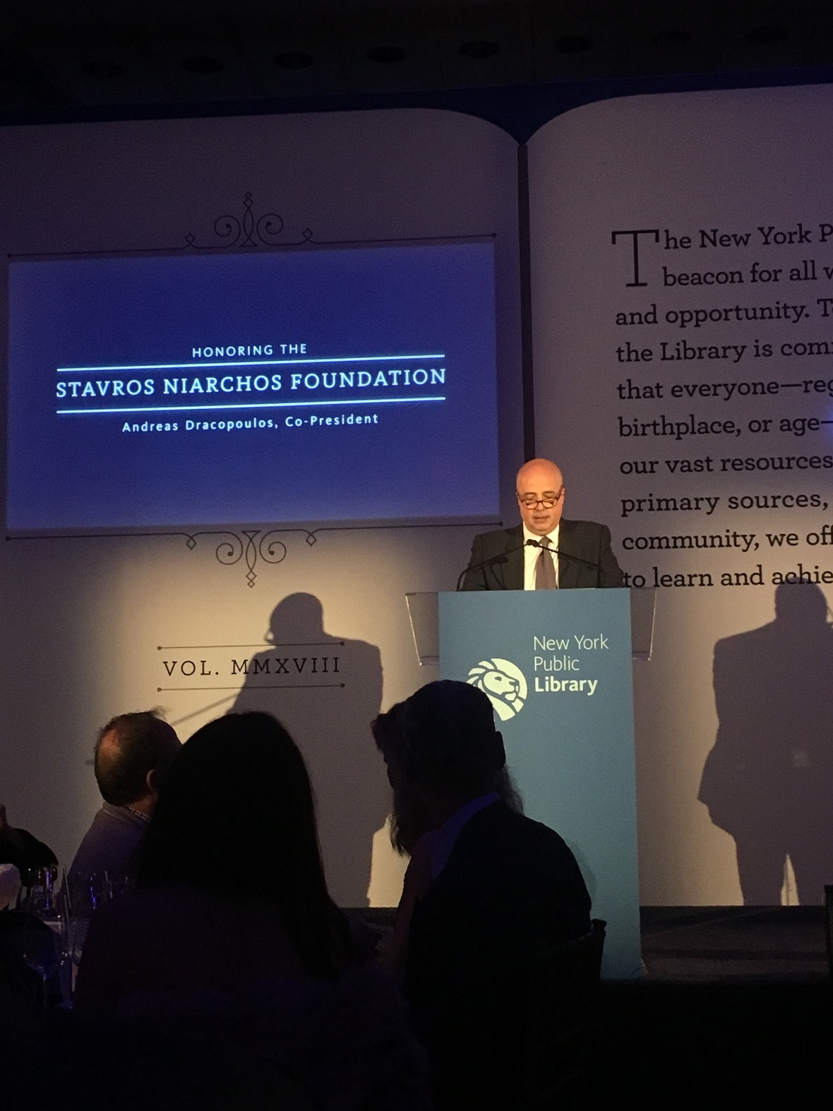 Ο Ανδρέας Δρακόπουλος, πρόεδρος του Ιδρύματος Σταύρος Νιάρχος, εκφωνεί ομιλία στο γκαλά της Δημόσιας Βιβλιοθήκης της Νέας Υόρκης, μπροστά από μια οθόνη που γράφει: 
