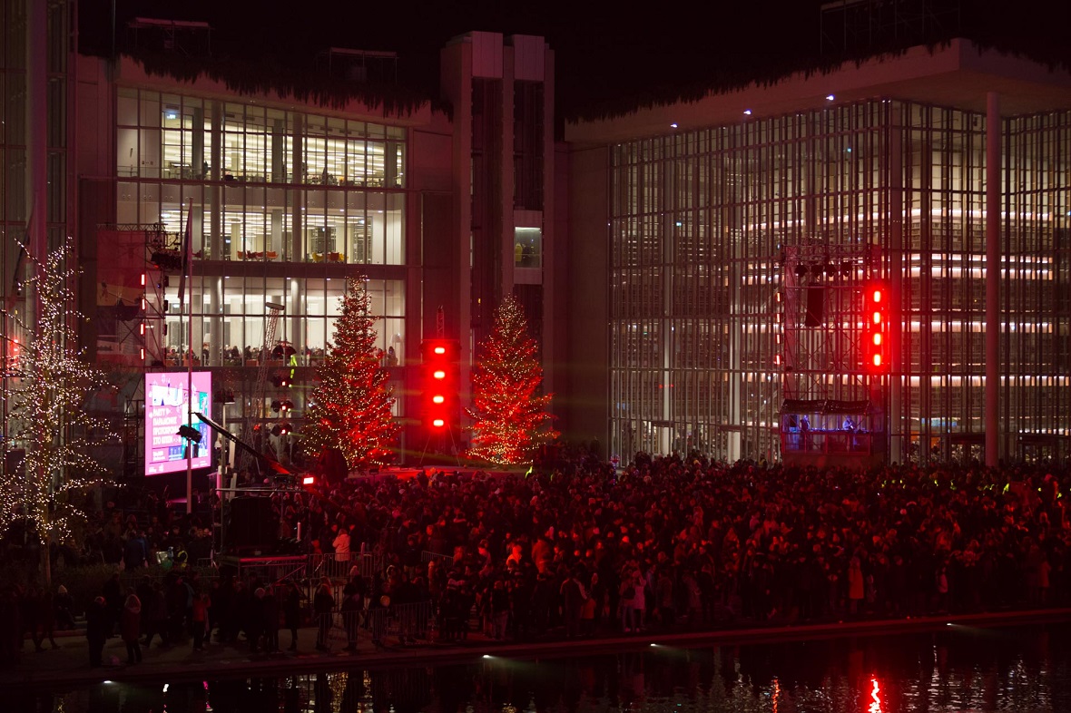 Μακρινή φωτογραφία που δείχνει πλήγθος κόσμου να παρακολουθεί μια συναυλία σε εξωτερικό χώρο δίπλα από δύο μεγάλα γυάλινα κτήρια