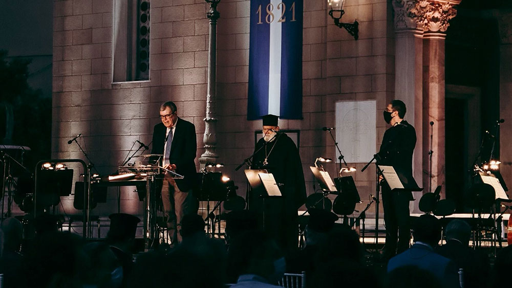 Σε μια σκηνή βρίσκονται τρεις άντρες, ο ένας μιλάει στο βήμα, ο δεύτερος είναι ιερέας και στέκεται ενώ ο τρίτος τους παρακολουθεί 