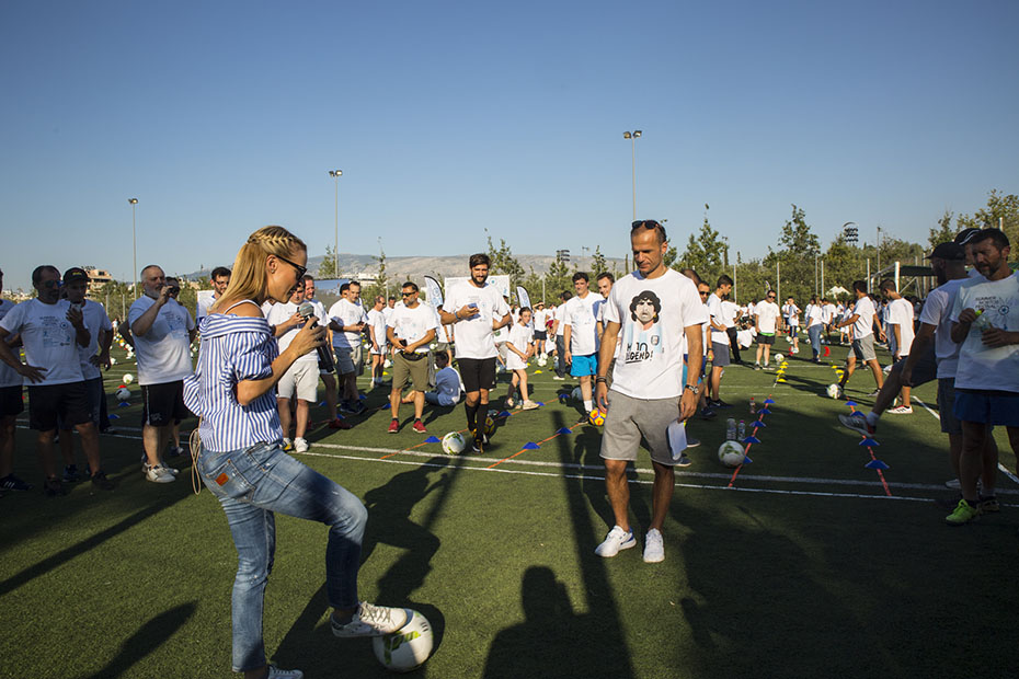 Μια γυναίκα κρατάει ένα μικρόφωνο και μιλάει και ταυτόχρονα έχει στα πόδια της μια μπάλα ποδοσφαίρου και γύρω της ο κόσμος την παρακολουθεί 