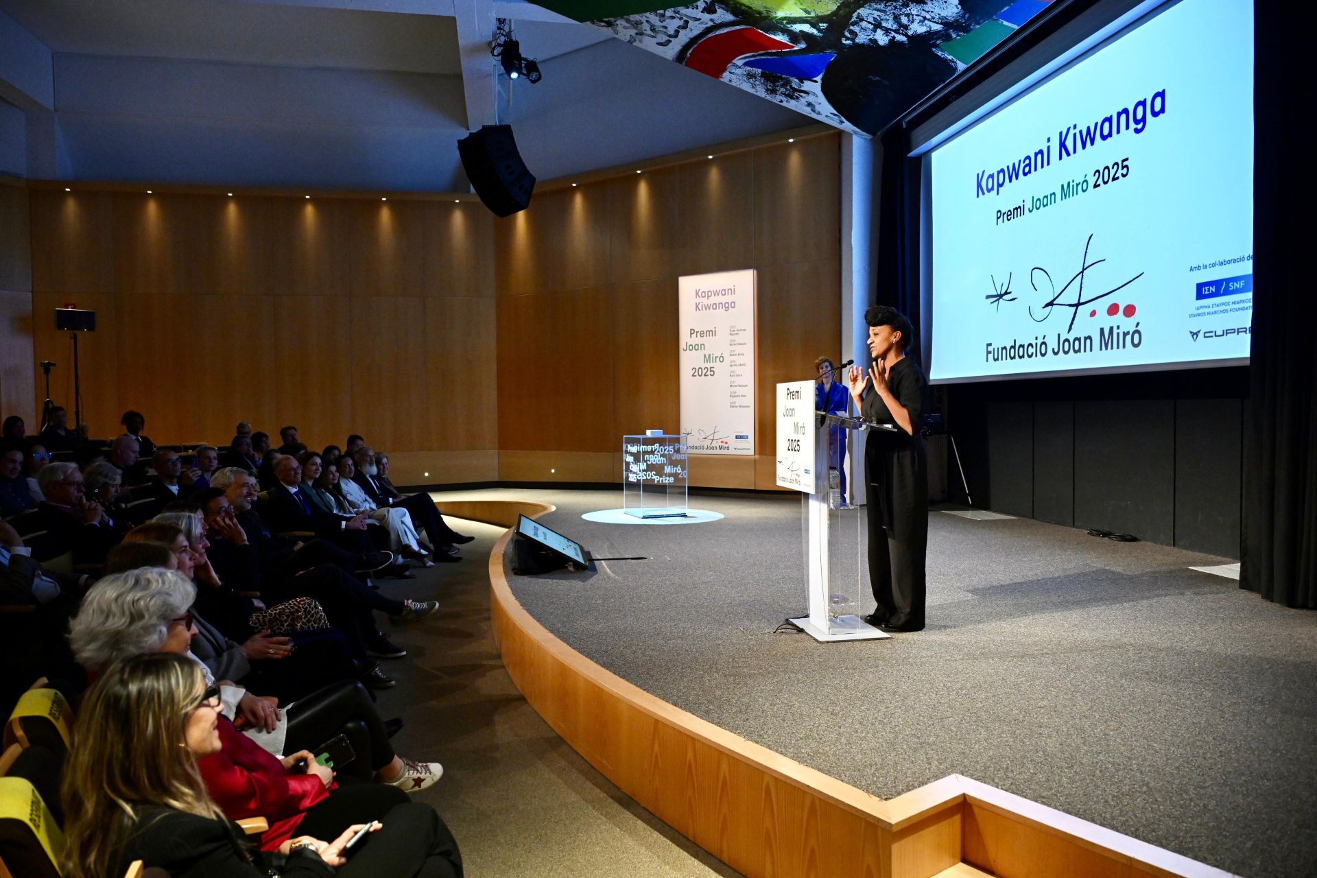 A woman on stage speaks at the podium during Joan Miró Prize 2025 ceremony, addressing an attentive audience seated in an auditorium and a large screen behind displays logos and her name, Kapwani Kiwange.