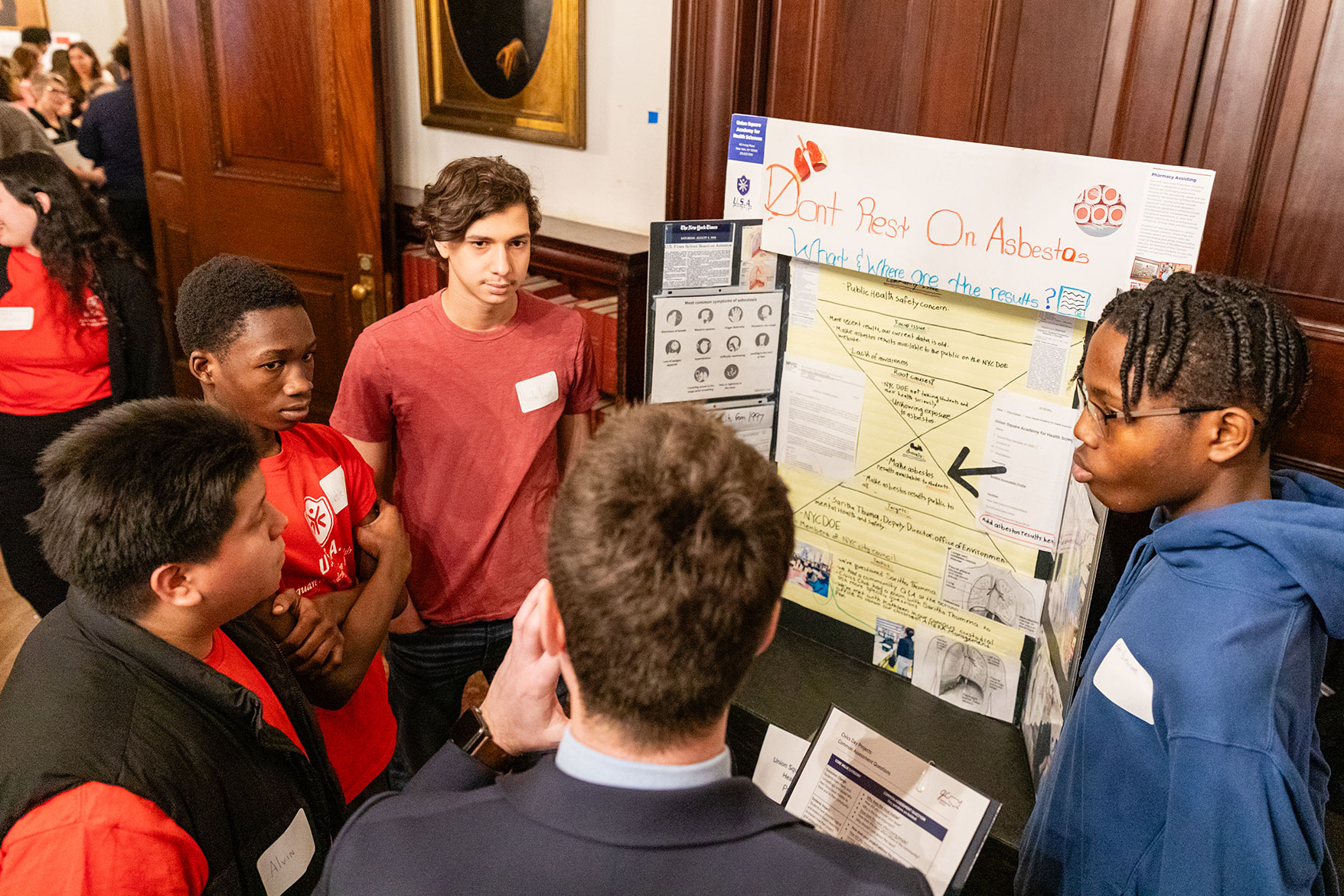 A group of high school students in conversation with an adult stand in front of a presentation posterboard titled “Don’t Rest on Asbestos.”