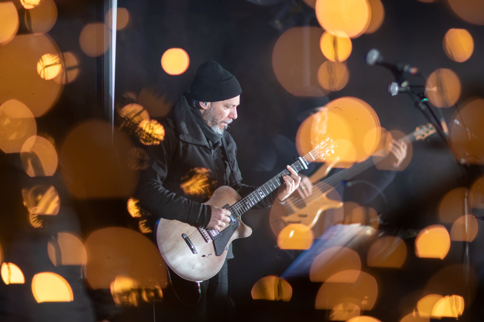 Ο Αλκίνοος Ιωαννίδης παίζει κιθάρα σε μια σκηνή που περιβάλλεται από Χριστουγεννιάτικα φώτα.