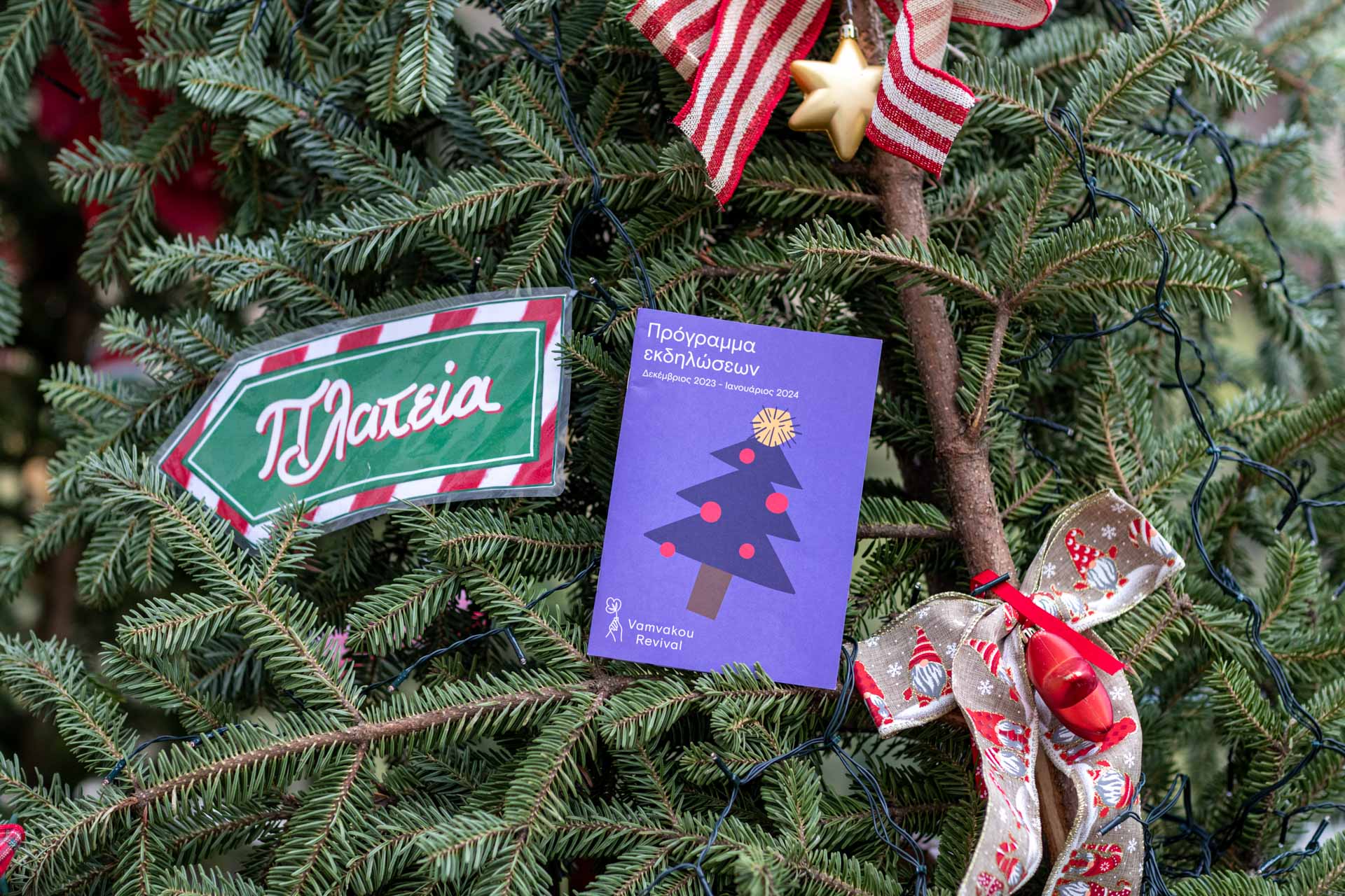 Το πρόγραμμα εκδηλώσεων είναι τοποθετημένο πάνω σε ένα χριστουγεννιάτικο έλατο και δίπλα του υπάρχουν χριστουγεννιάτικοι φιόγκοι καθώς και ένα βελάκι το οποίο γράφει πλατεία δείχνοντας την κατεύθυνση προς αυτή. 