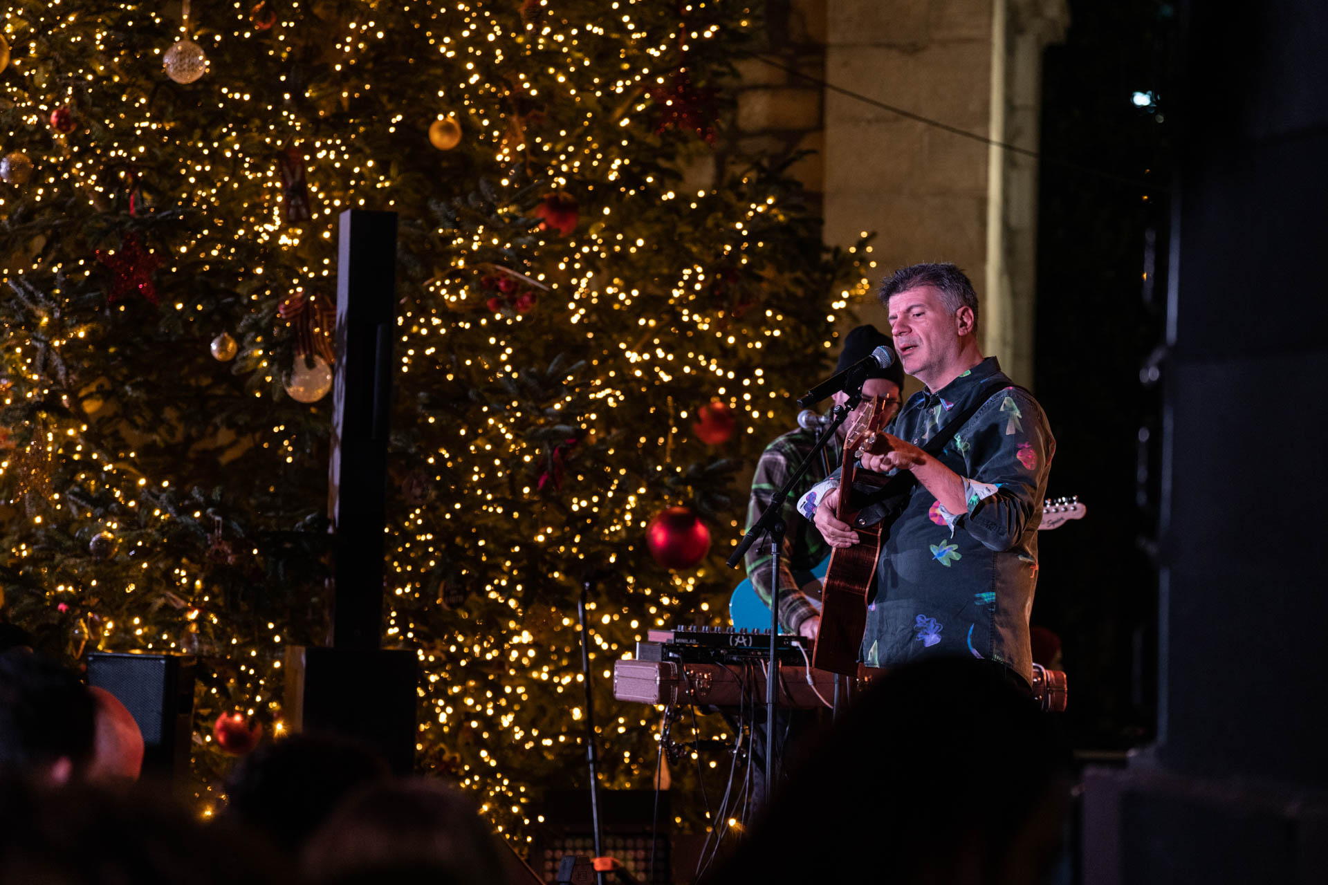 Ο Φοίβος Δεληβοριάς είναι πάνω στη σκηνή παίζει κιθάρα και τραγουδάει ενώ γύρω του υπάρχουν στολισμένα χριστουγεννιάτικα δέντρα και το κοινό τον παρακολουθεί.