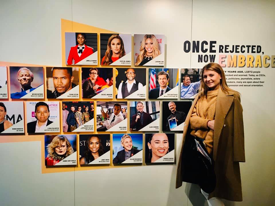 Μια γυναίκα στέκεται δίπλα σε έναν τοίχο ο οποίος είναι διακοσμημένος με ένα κολάζ διάσημων προσώπων και φωτογραφίζεται 