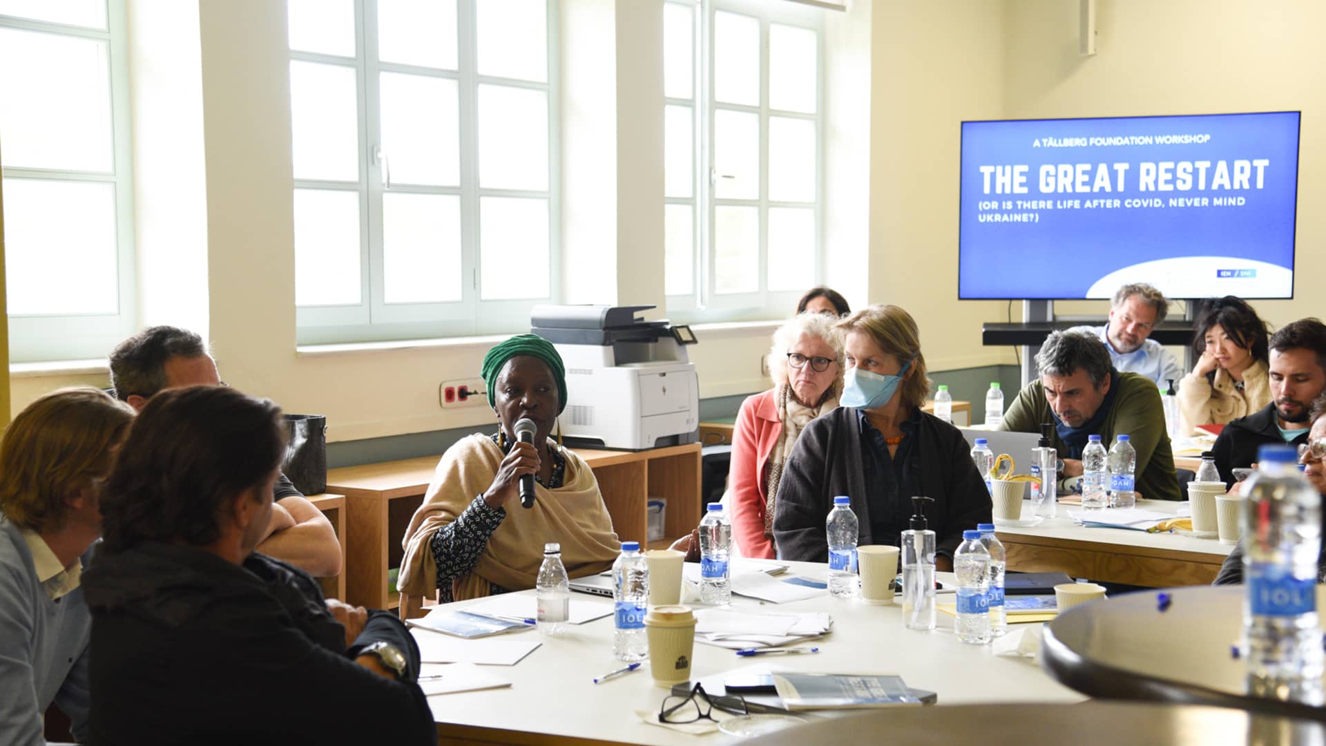 Μια ομάδα ανθρώπων κάθεται στο τραπέζι και ακούν μια κυρία που κρατάει ένα μικρόφωνο και τους μιλάει 