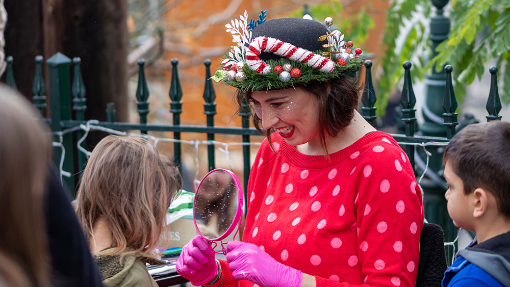 Μια γυναίκα ντυμένη με χριστουγεννιάτικη ενδυμασία κρατάει έναν καθρέφτη για να δει ένα παιδί το αποτέλεσμα του προσώπου του