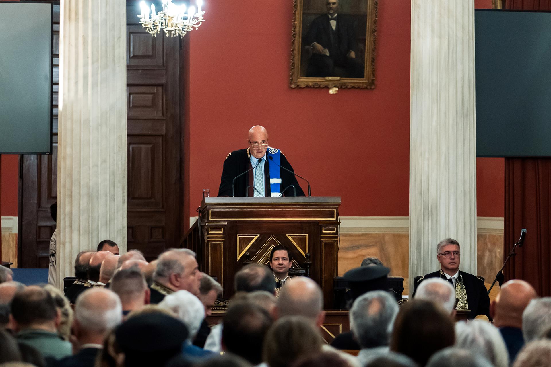 Μέλη από το κοινό παρακολουθούν τον τιμώμενο Πρόεδρο του ΙΣΝ, Ανδρέα Δρακόπουλο, καθώς εκφωνεί ομιλία από το βήμα της αίθουσας.