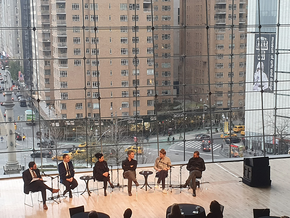 Πέντε άνθρωποι κάθονται και συζητούν μπροστά σε κοινό με φόντο την πόλη 